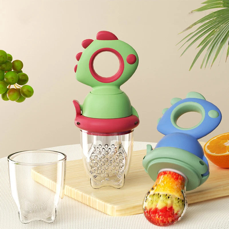 A green dinosaur rotating fruit feeder for babies is shown next to a glass and an orange on a wooden surface.