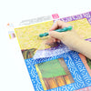 A hand is using a tool to release a green bead from a sheet of reusable non-stick release paper for diamond painting.