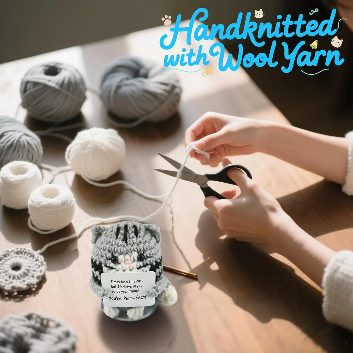 Hands are knitting a small gray and white crocheted cat figurine with wool yarn on a wooden table.