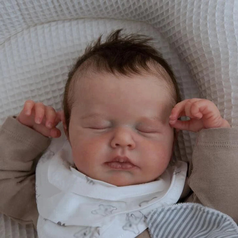 A newborn baby doll with rooted hair is sleeping, wrapped in a white bonnet and clothing.