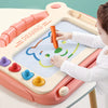 A child is drawing on a pink magnetic board with an orange marker and various colored magnetic shapes.