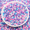 A white bowl filled with colorful polymer clay slice sprinkles in various shades of pink, blue, and purple scattered on a sur