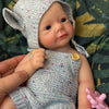 A hand-painted silicone reborn doll wearing a knitted hat and outfit, resting on green fabric.