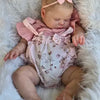 A newborn baby doll with hand-painted skin wearing a pink outfit and bow, resting in white fluffy material.