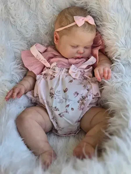 A newborn baby doll with hand-painted skin wearing a pink outfit and bow, resting in white fluffy material.