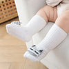 A pair of white cotton socks with gray cartoon animal patterns are shown on a baby's foot resting on a white sofa.