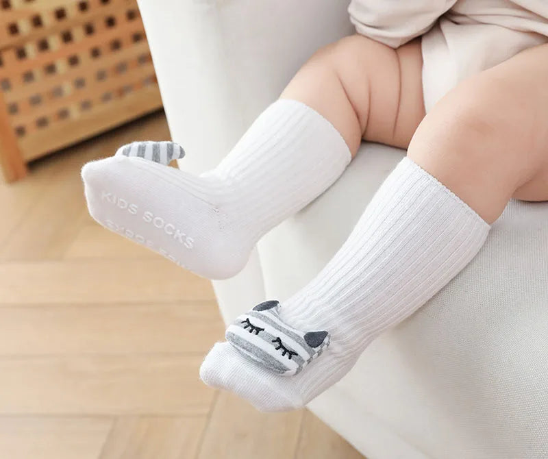 A pair of white cotton socks with gray cartoon animal patterns are shown on a baby's foot resting on a white sofa.