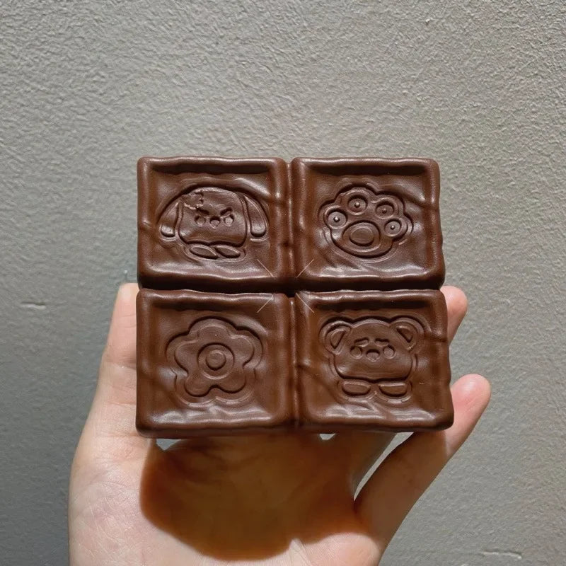 A hand holds a square brown squishy toy with four embossed designs on it.