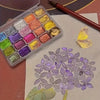 A watercolor paint set in a clear travel tin with various shades of iridescent and jewel-toned colors, next to a paintbrush a