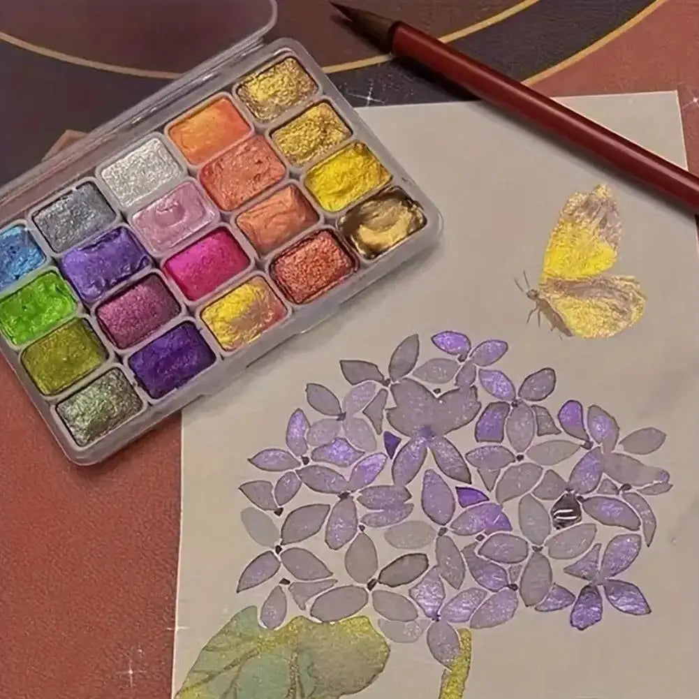 A watercolor paint set in a clear travel tin with various shades of iridescent and jewel-toned colors, next to a paintbrush a