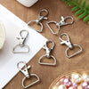 A collection of silver swivel snap hooks with D-rings for keychains and craft projects arranged on a brown fabric surface.