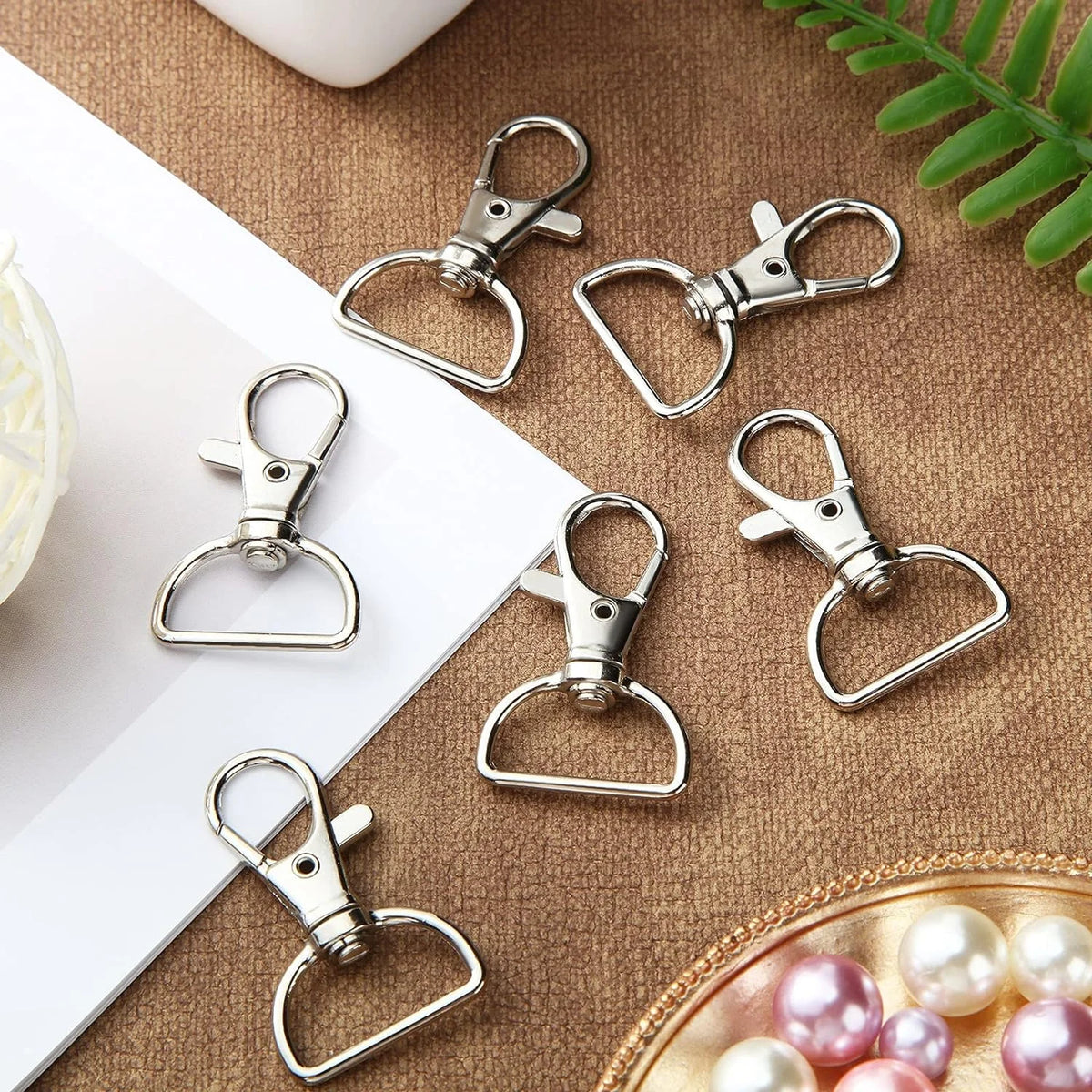 A collection of silver swivel snap hooks with D-rings for keychains and craft projects arranged on a brown fabric surface.