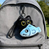A cute plush black cat keychain with yellow eyes and a light blue fish charm attached to a gray backpack.