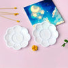A set of ten flower-shaped white disposable paint palettes rests on a pink surface next to a blue celestial painting.