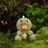 A soft green plush doll resembling a creature with large eyes and horns sitting in a lush, green outdoor setting.