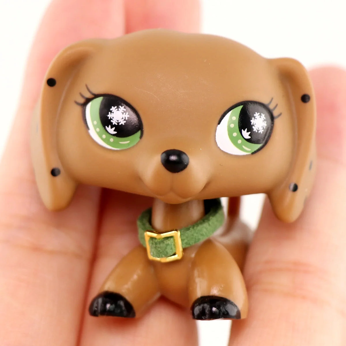 A small, brown miniature pet animal figure with large green eyes and a green collar is held in a person's hand.