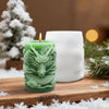 A green silicone candle mold shaped like a dragon with cloud patterns sits on a wooden surface with pine needles and snow.