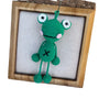 Handmade crochet frog hanging ornament with large eyes and a cross symbol displayed in a wooden frame