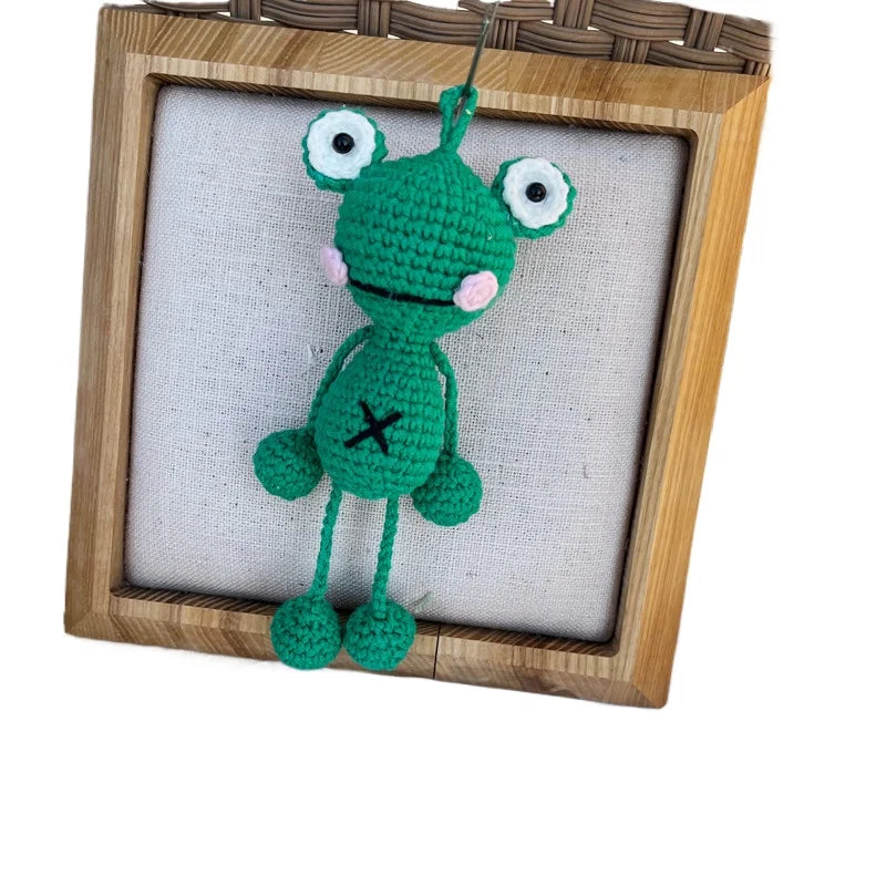 Handmade crochet frog hanging ornament with large eyes and a cross symbol displayed in a wooden frame