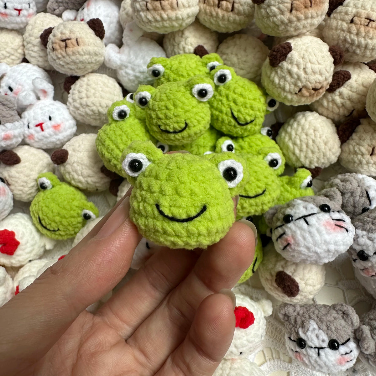 A hand holds a bright green crocheted animal keychain among many similar small stuffed animals.