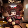 A brown bear plush charm product featuring a dark brown fur and reddish-brown clothing, displayed among cookies.