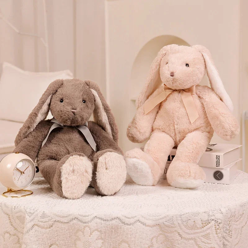 A soft, light pink bunny plush toy with long ears and a bow, sitting next to a small clock.