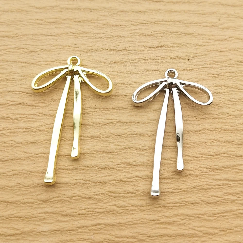 A pair of gold-toned alloy bow charms for jewelry making displayed on a neutral brown surface.