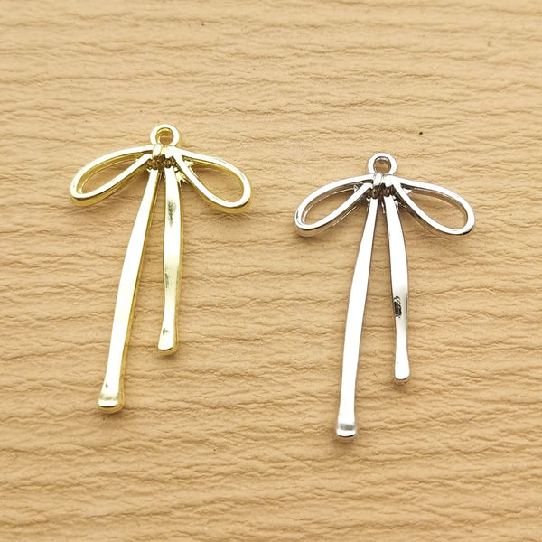 A pair of gold-toned alloy bow charms for jewelry making displayed on a neutral brown surface.