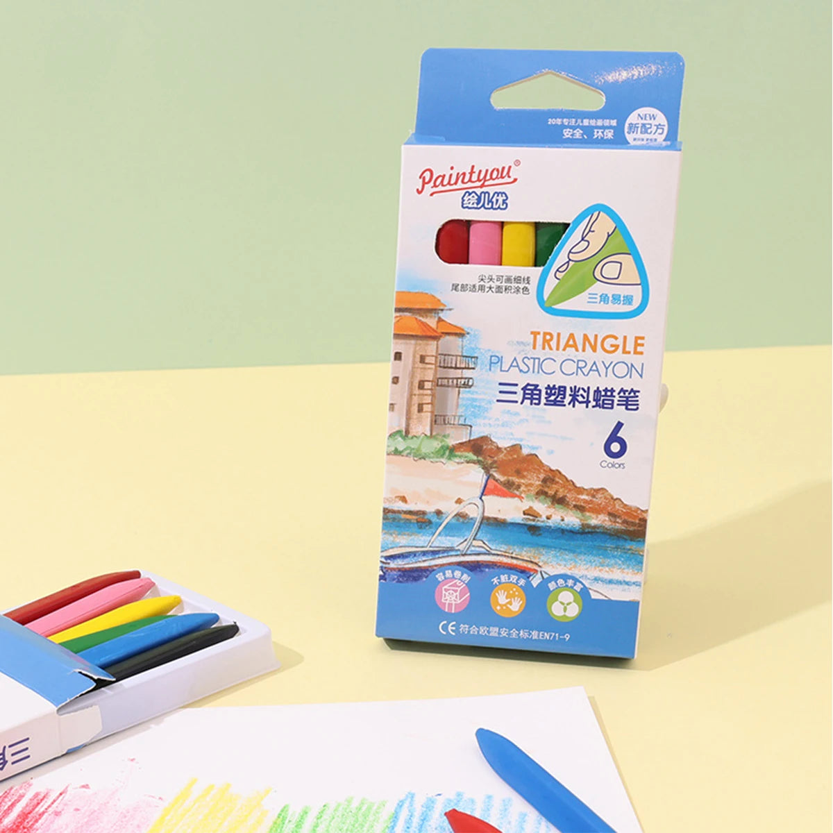 A box of six triangle plastic crayons with a green and blue tip is shown next to a white paper and a blue crayon.