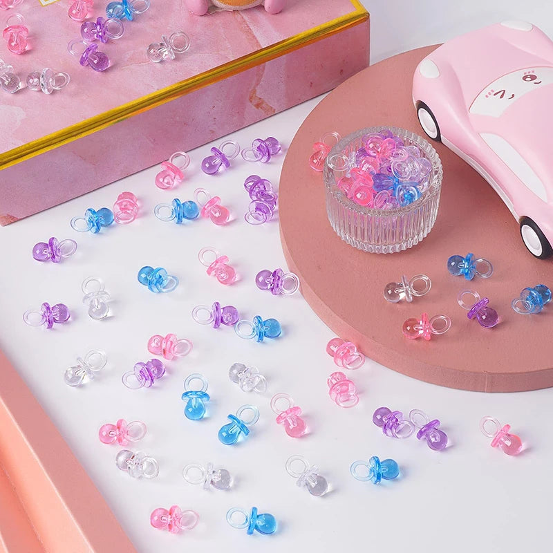 A collection of mini acrylic pacifier party favors in various shades of pink, purple, and blue are displayed on a white surfa