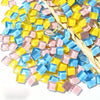 A pile of small, colorful crystal glass mosaic tiles in shades of yellow, blue, and pink resting on straw.