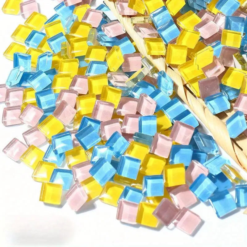 A pile of small, colorful crystal glass mosaic tiles in shades of yellow, blue, and pink resting on straw.