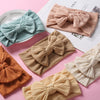 Three soft stretch baby headbands with bows in shades of teal, beige, and orange are displayed on a pink background.
