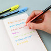A hand writes in a notebook using a neon colored pen next to several metallic glitter gel pens.