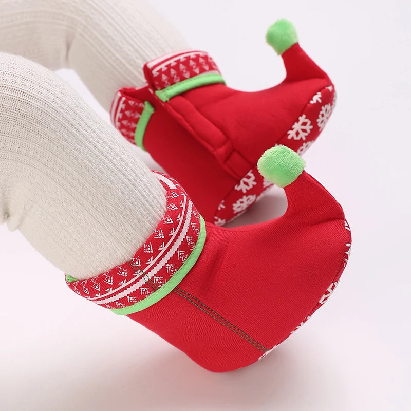 A pair of red baby first walker shoes with green accents and white snowflake patterns, shown on a person's foot.