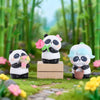 A blind box panda collectible figure with black and white fur and pink accents is displayed on a wooden block amidst green fo