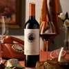 A bottle of red wine with a natural wood tapered bottle and test tube stopper is displayed on a wooden table.