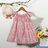 A pink floral smocked summer dress for girls with ruffled sleeves hanging on a wooden hanger.