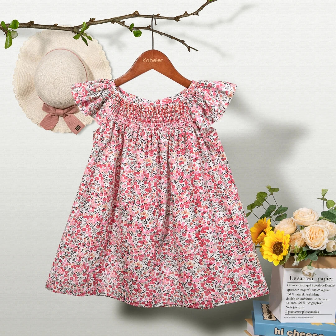 A pink floral smocked summer dress for girls with ruffled sleeves hanging on a wooden hanger.