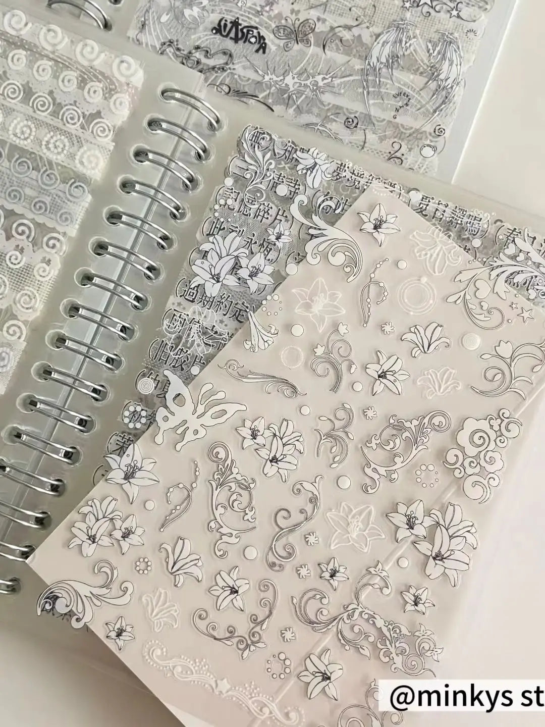 A close-up of a sticker book with 32 sheets featuring delicate gray and white floral and decorative designs for journaling.