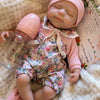 A newborn silicone reborn baby doll wearing a floral outfit is resting on a fluffy white blanket.