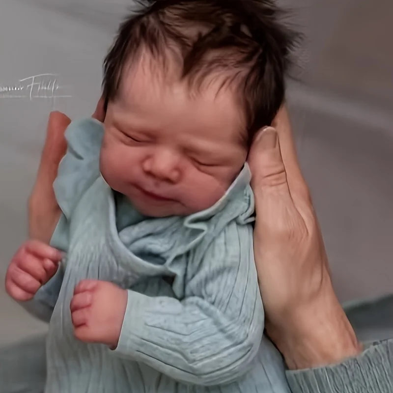A newborn baby doll with soft cloth body wearing a light blue knitted outfit, held by a person's hands.