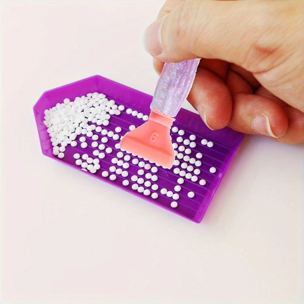 A hand is using a tool to apply white beads onto a purple diamond painting canvas.