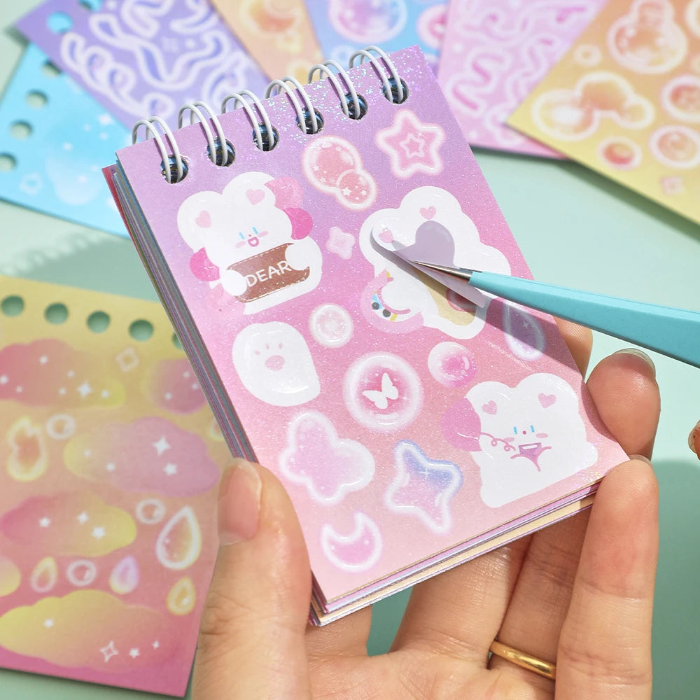 A hand holds a pink aesthetic sticker book with various pastel designs and a blue pen against a light green background.