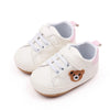 A pair of cream-colored baby sneakers with pink accents and a brown bear face detail.