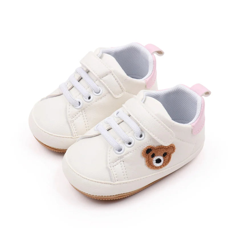 A pair of cream-colored baby sneakers with pink accents and a brown bear face detail.