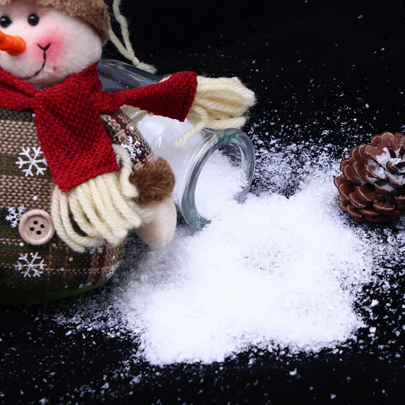 A close-up of artificial snow powder scattered on a black surface next to a knitted doll and a pinecone.