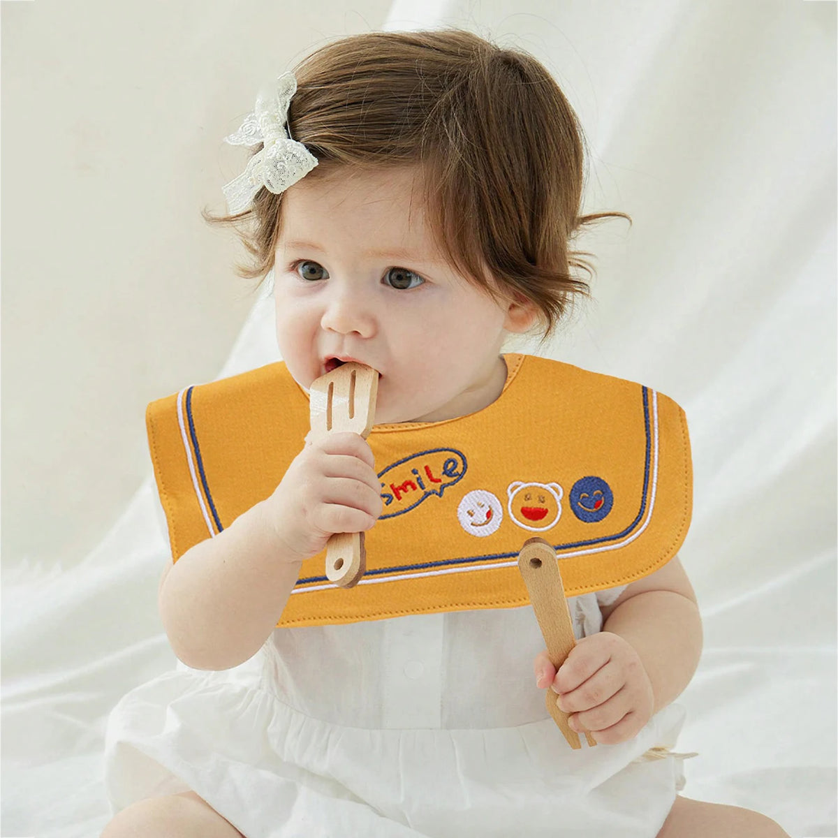 A baby is holding a yellow bib with a smile design, wearing a white outfit.