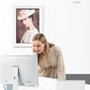 A woman is looking at a framed photo while working on a computer and a laptop are visible on a white desk.