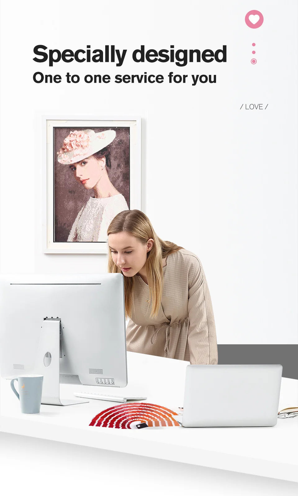 A woman is looking at a framed photo while working on a computer and a laptop are visible on a white desk.